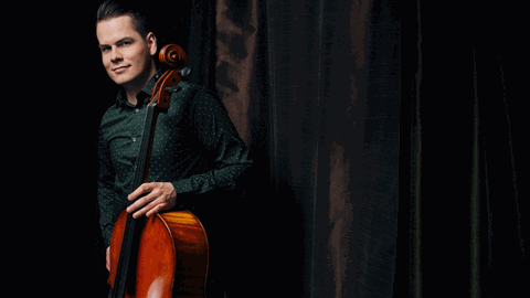 Ein junger Mann mit gegeelten Haaren steht in dunkelgrünem Hemd neben einem dunklen Vorhang mit dem aufgestellten Cello in der Hand, sodass die Schnecke des Instrumentes neben seinem Ohr erscheint.