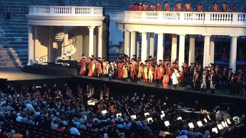 Zu sehen ist die große Freiluftbühne der Arena di Verona in der gleichnamigen italienischen Stadt. Auf der Bühne findet eine Opernaufführung statt: der Maskenball von Guiseppe Verdi.