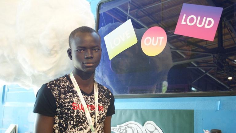 Der Blogger John Bjor Ajang aus dem Südsudan steht for einer mit Aufklebern beklebten Wand, er trägt ein t-Shirt und schaut in die Kamera
