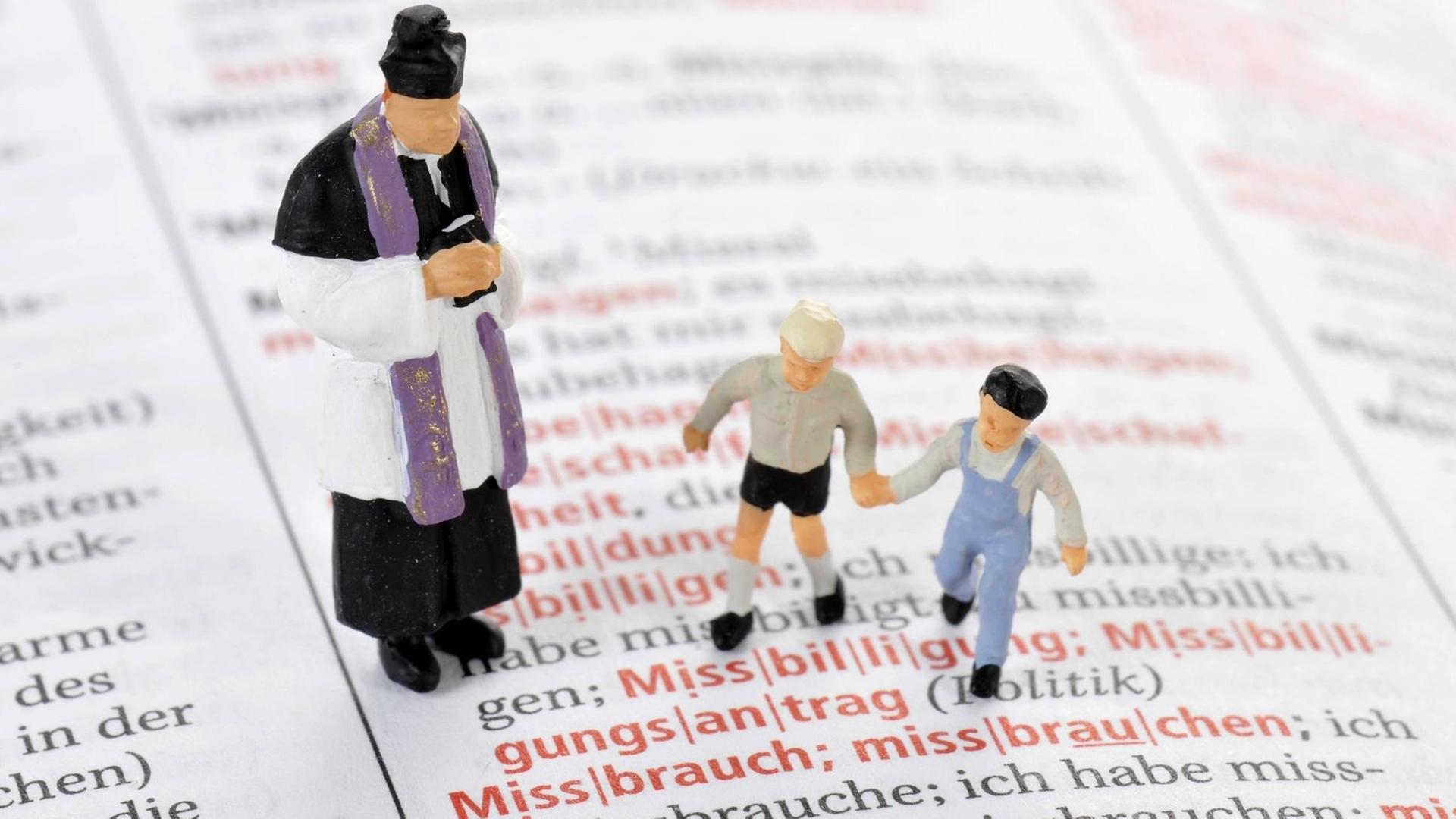 Modelleisenbahnfiguren eines Priesters und zweier Kinder auf einer aufgeschlagenen Wörterbuchseite vor dem Wort "Missbrauch"