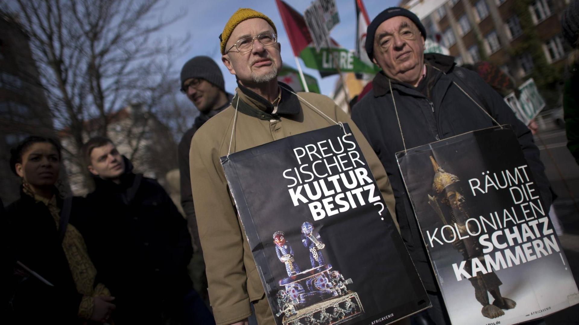 Demonstranten auf dem Gedenkmarsch zur deutschen Kolonialgeschichte mit Plakaten um den Hals, wo der Preussische Kulturbesitz hinterfragt wird und aufgefordert wird, die kolonialen Schatzkammern zu räumen.