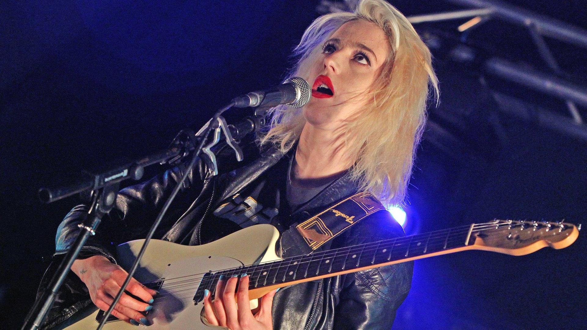 Du Blonde 2012 noch als Beth Jeans Houghton auf dem Applecart Festival in London. Blonde Sängerin, rote Lippen, schwarze Lederjacke, schwarz-weiße Hose, eine E-Gitarre spielend und singend auf einer Bühne