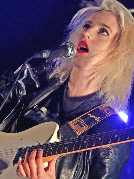 Du Blonde 2012 noch als Beth Jeans Houghton auf dem Applecart Festival in London. Blonde Sängerin, rote Lippen, schwarze Lederjacke, schwarz-weiße Hose, eine E-Gitarre spielend und singend auf einer Bühne