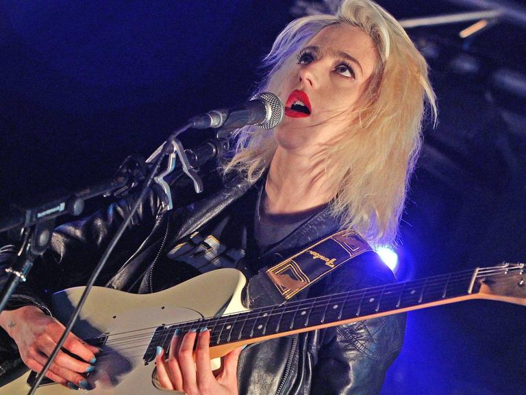 Du Blonde 2012 noch als Beth Jeans Houghton auf dem Applecart Festival in London. Blonde Sängerin, rote Lippen, schwarze Lederjacke, schwarz-weiße Hose, eine E-Gitarre spielend und singend auf einer Bühne