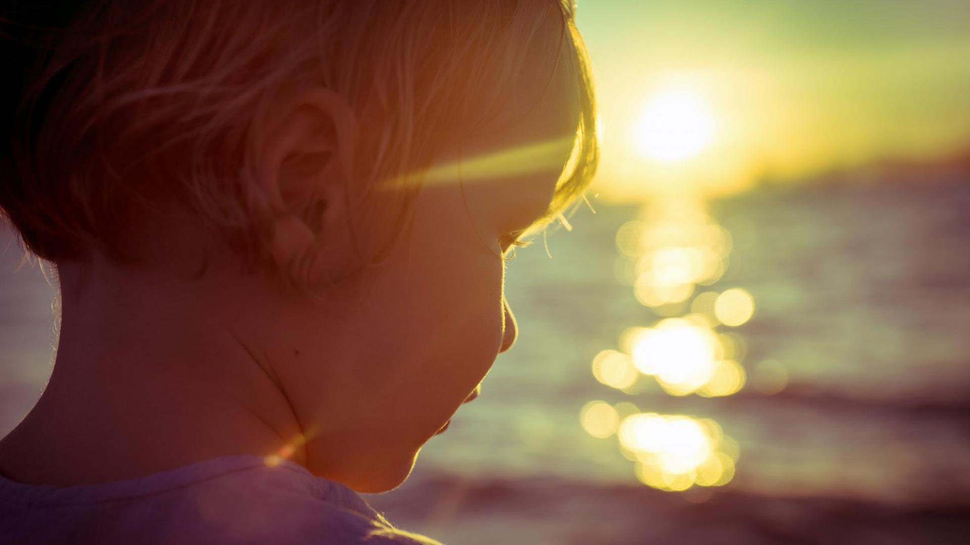 Fotografie eines Kindes am Meer, im Gegenlicht der, am Horizont, untergehenden Sonne.