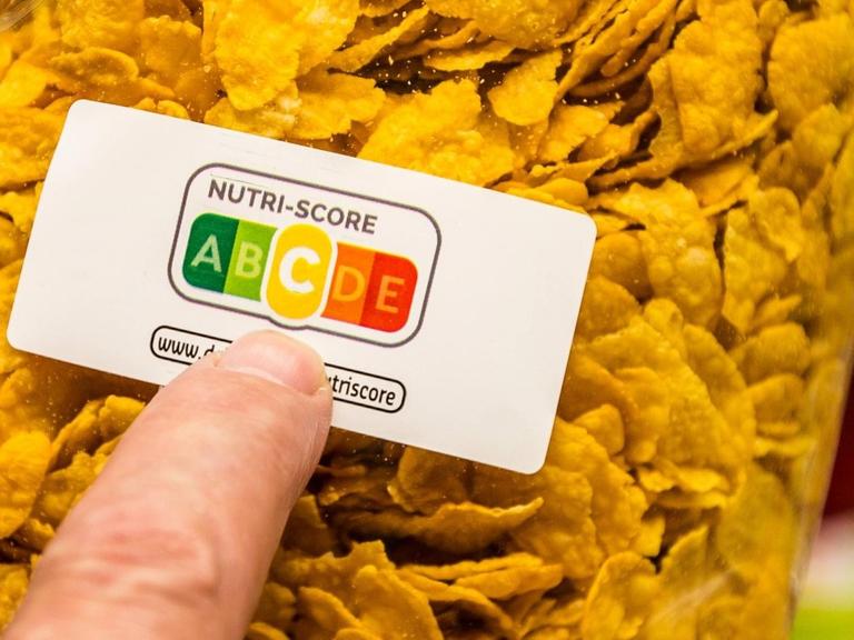 Eine Packung Cornflakes, ein Zeigefinger deutet auf die Farbflächen des Nutri-Score-Aufdrucks. Die mittlere Kategorie "C" (gelb) ist hervorgehoben.
