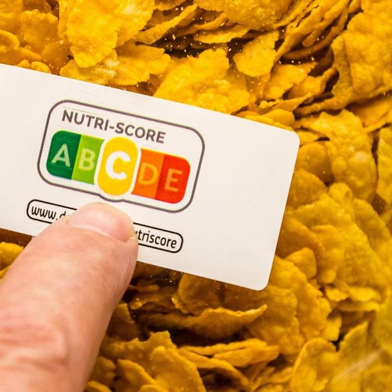 Eine Packung Cornflakes, ein Zeigefinger deutet auf die Farbflächen des Nutri-Score-Aufdrucks. Die mittlere Kategorie "C" (gelb) ist hervorgehoben.