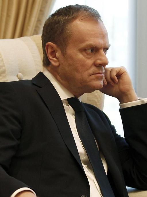 Tusk sitzt in einem beigen Sessel vor einem Fenster mit weißem Vorhang und hat eine Hand an das Kinn gestützt. Er blickt konzentriert und scheint jemandem aufmerksam zuzuhören.
