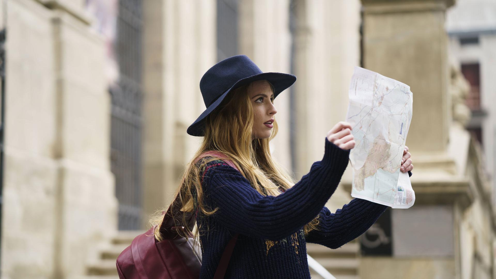 Eine junge Frau mit Hut und Rucksack steht in einer Straße und schaut auf einen Stadtplan.