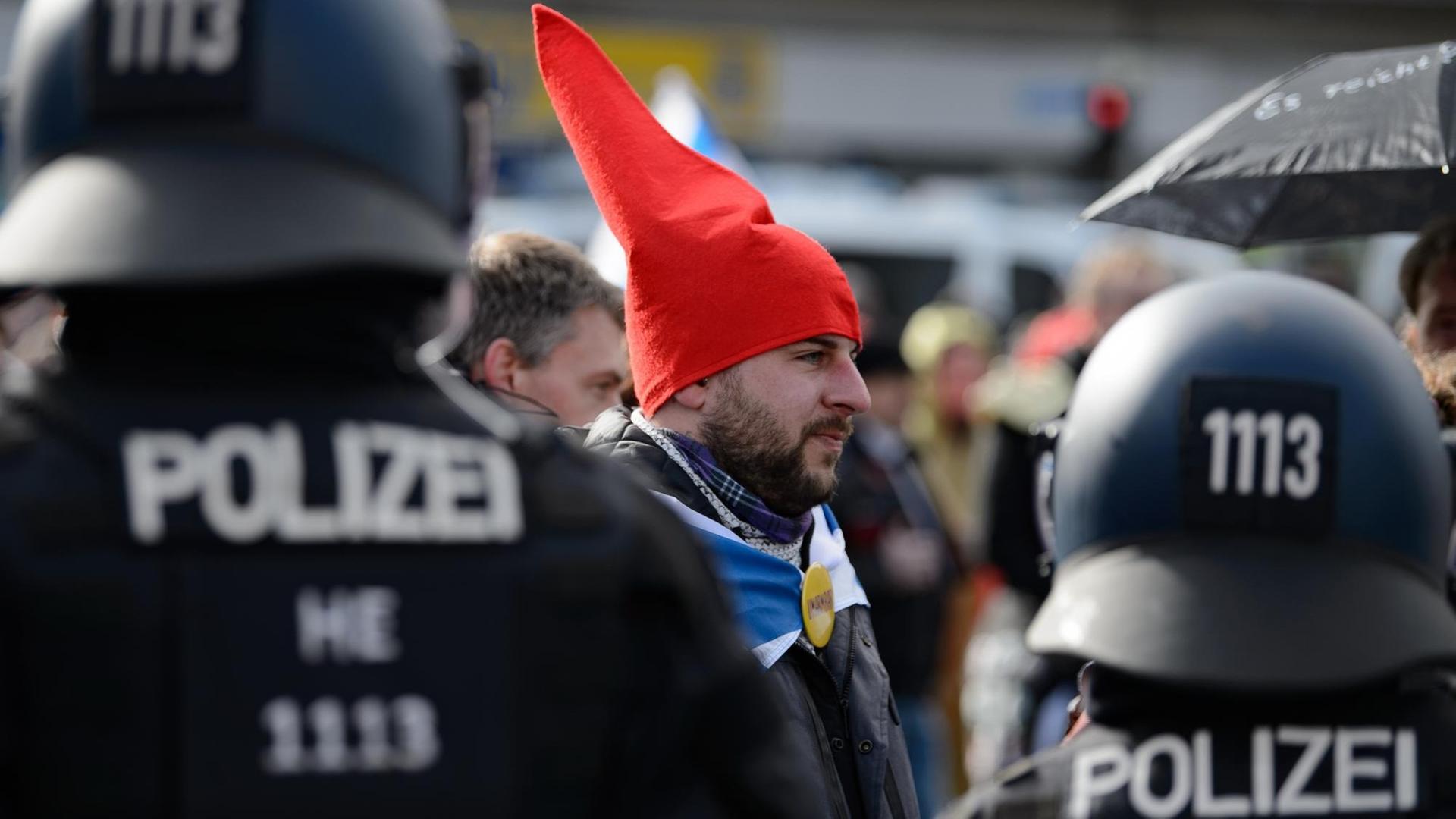 Ein Teilnehmer mit einer roten Zipfelmütze und ohen Mund-Nasen-Bedeckung steht bei einer Kundgebung unter dem Motto "Freie Bürger Kassel - Grundrechte und Demokratie" vor Polizisten.