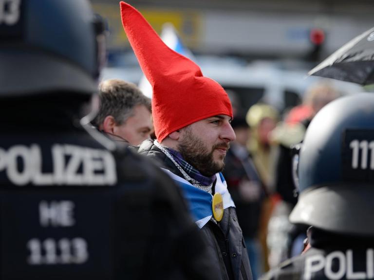 Ein Teilnehmer mit einer roten Zipfelmütze und ohen Mund-Nasen-Bedeckung steht bei einer Kundgebung unter dem Motto "Freie Bürger Kassel - Grundrechte und Demokratie" vor Polizisten.