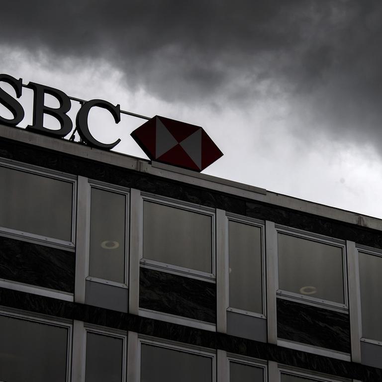 Schriftzug HSBC auf einem Gebäude, über dem dunkle Wolken aufziehen.