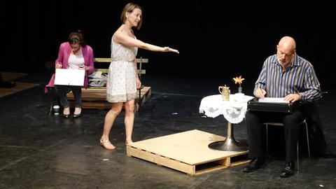 Eine Theaterbühne: Links sitzt eine Frau auf einer Bank und schreibt, in der Mitte steht eine Frau und macht eine anschuldigende Geste in Richtung eines Mannes, der rechts von ihr sitzt und schreibt.
