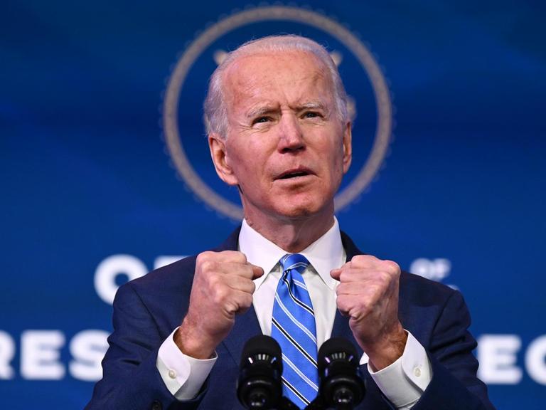 Joe Biden hat während einer Rede auf einem Podium beide Hände erhoben.