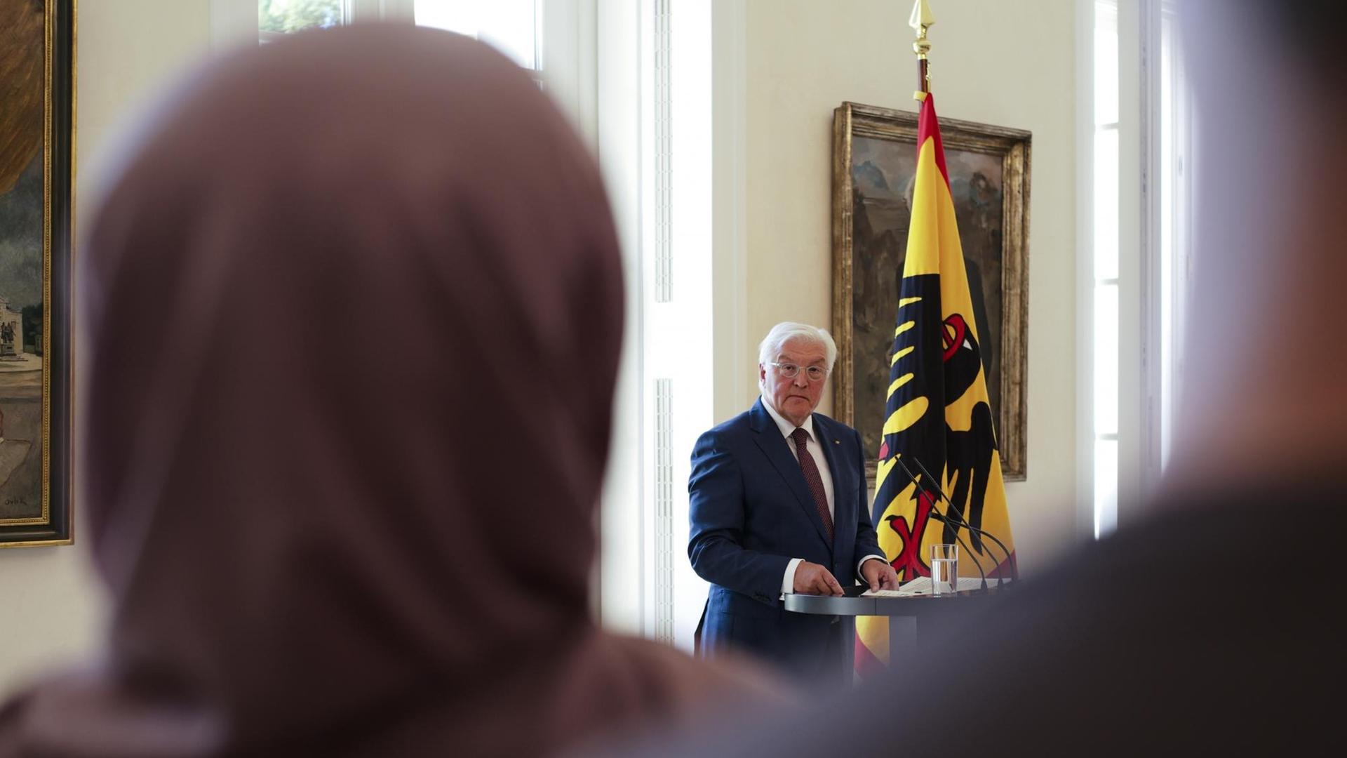 Bundespräsident Steinmeier steht am Rednerpult, im Vordergrund des Bildes ist eine Frau mit Kopftuch von hinten zu sehen.