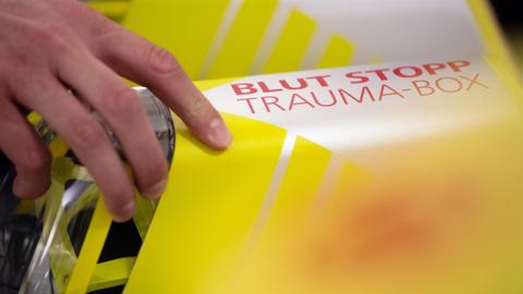 Eine Hand greift nach der Trauma-Box mit der Aufschrift «Blut Stopp». Die Box enthält ein sogenanntes Tourniquet, ein Abbindesystem ähnlich einer Blutdruckmanschette, und einen saugfähigen Druckverband zum Stillen einer Blutung eines Verletzten, bis medizinisches Fachpersonal eintrifft.