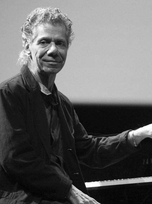 Ein Mann mit grauen Locken und dunklem Hemd sitzt auf einer Bühne an einem Flügel, von dessen Tastatur er sich abwendet, um Richtung Publikum zu schauen.