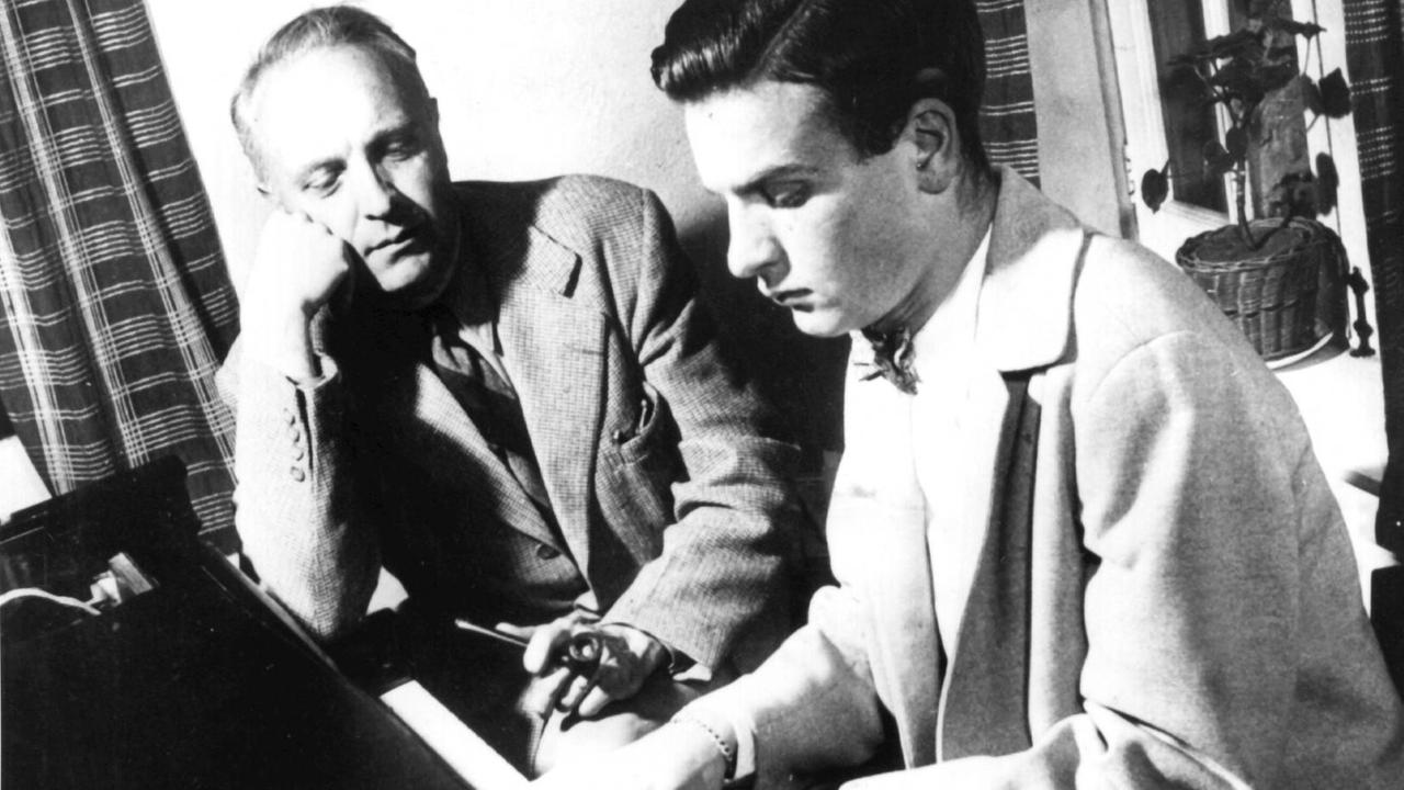 Schwarz-Weiß Aufnahme von 1956 mit Robert (li) und Jean Casadesus (re), der an einem schwarzen Flügel sitzt und spielt, sein Vater hört ihm zu, der rechte Arm ist aufgestützt, in der linken hält er eine Pfeife, beide tragen Anzüge