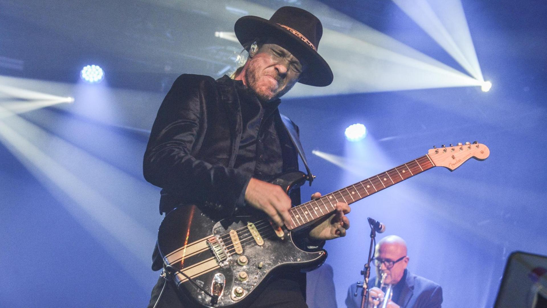 Ein Mann mit schwarzem Samt-Jackett, blonden Haaren und Hut auf dem Kopf steht im Scheinwerferlicht auf der Bühne und spielt mit geschlossenen Augen eine schwarze E-Gitarre.