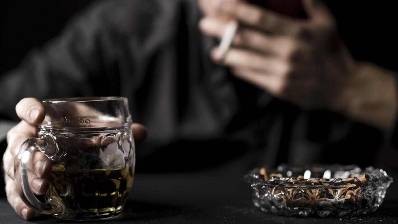 Ein Glas Alkohol und ein voller Aschenbecher, dazu unscharf im Hintergrund ein Mann, der das Glas in der Hand hält und an einer Zigarette zieht. (Symbolbild)