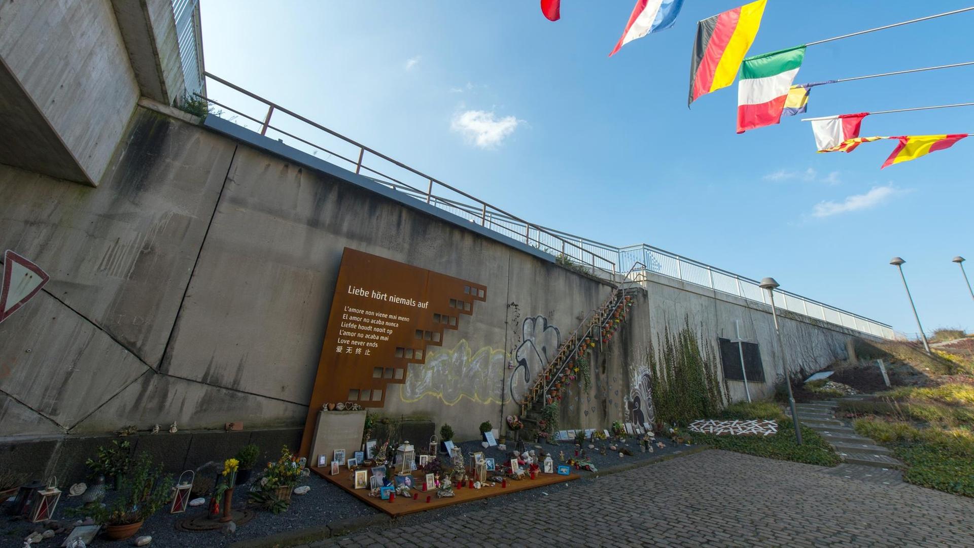 Eine langgezogene Mauer mit einer Gedenktafel, davor Blumen und Fotos.