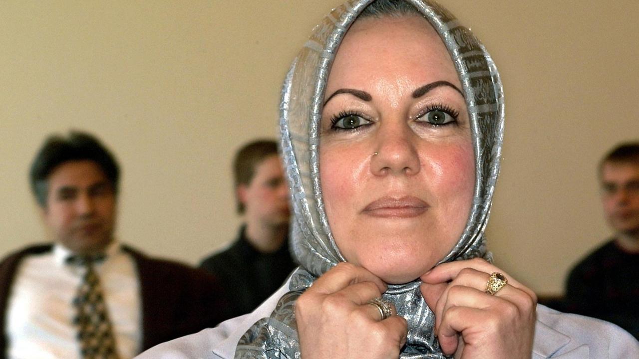 Die niedersächsische Lehrerein Iyman Alzayed zupft an ihrem silberfarben-glänzenden Kopftuch. Hinter ihr sieht man im Gerichtssaal unscharf drei Männer sitzen.