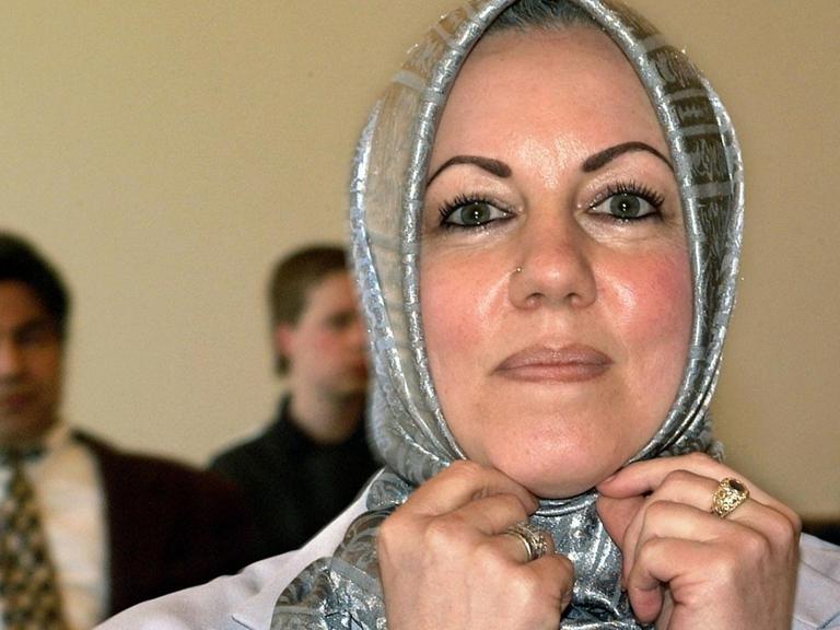 Die niedersächsische Lehrerein Iyman Alzayed zupft an ihrem silberfarben-glänzenden Kopftuch. Hinter ihr sieht man im Gerichtssaal unscharf drei Männer sitzen.