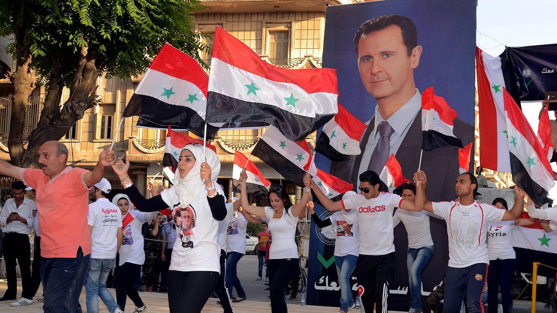 Junge Männer und Frauen mit Kopftüchern tragen syrische Flaggen und große Plakate mit dem Konterfei von Baschar al-Assad.
