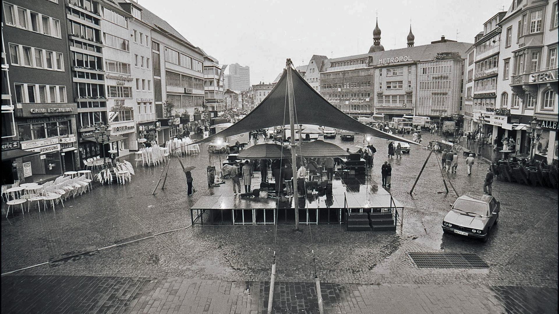 Eine Schwarzweißaufnahme von einem Markplatz in Bonn in der Wendezeit. Eine Bühne ist aufgebaut, wenige Menschen stehen davor. Sie halten Regenschirme, denn es regnet.