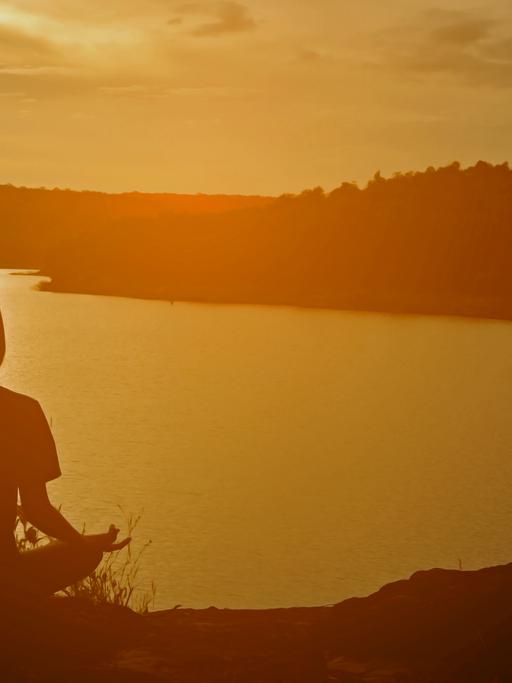 Eine Frau in Meditationshaltung sitzt in der Abendsonne auf einem Hügel mit Blick auf einen See.