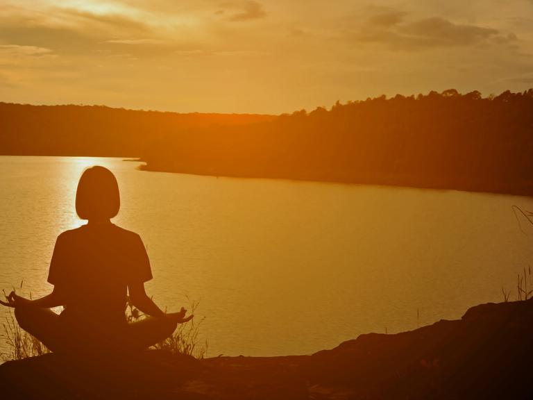 Eine Frau in Meditationshaltung sitzt in der Abendsonne auf einem Hügel mit Blick auf einen See.