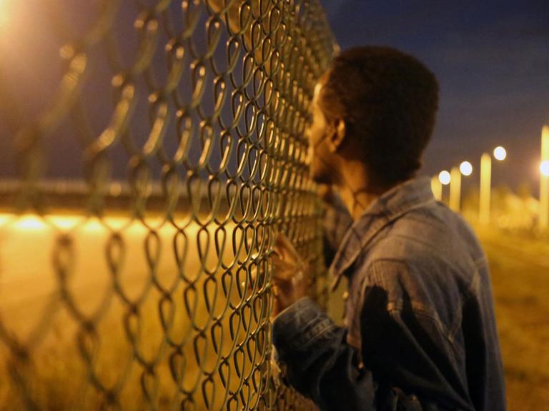 Ein Migrant schaut durch einen Zaun an den befestigten Anlagen am Eurotunnel nahe Calais