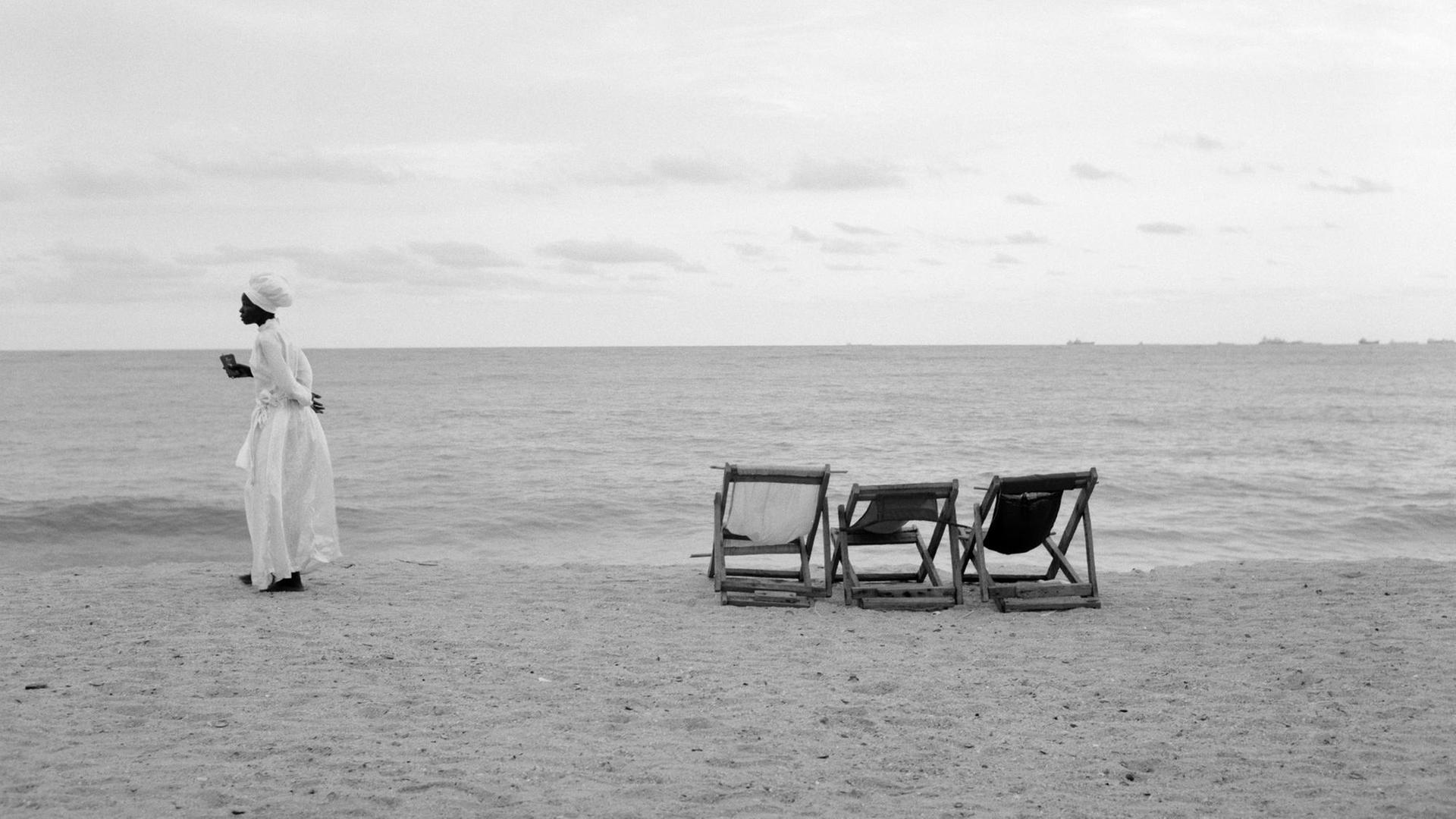 Eine Frau in weißem Kleid steht an einem Strand, neben ihr sind drei Standliegen.