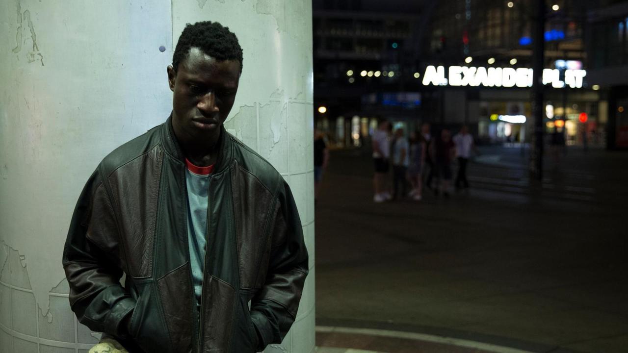 Der Schauspieler Welket Bungué steht im Film "Berlin Alexanderplatz" vor einer Säule, im Hintergrund der Neon-Schriftzug Alexanderplatz.