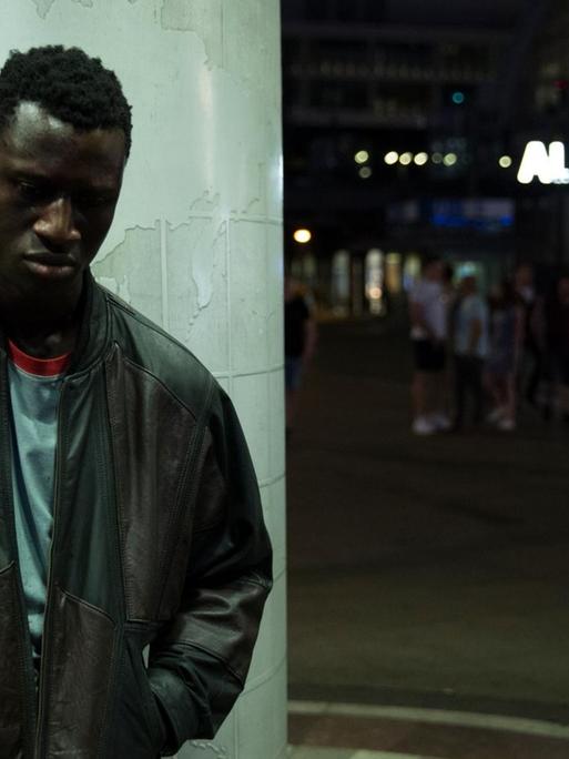 Der Schauspieler Welket Bungué steht im Film "Berlin Alexanderplatz" vor einer Säule, im Hintergrund der Neon-Schriftzug Alexanderplatz.