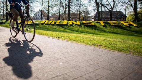 Eine Radfahrerin fährt auf der Promenade entlang, im Hintergrund steht der Schriftzug «Münster», aus Blumen gefertigt. Der ADFC-Fahrradklima-Test 2018 ist eine Befragung mit dem Ziel der vergleichbaren Erfassung von Radfahrbedingungen in Staädten und Gemeinden. Die Datenerhebung zum ADFC-Fahrradklima-Test 2018 erfolgte vom 1.9.2018 bis zum 30.11.2018. Deutschlandweit wurden Radfahrer gebeten, ihre Einschätzung zum Radverkehr in ihrer Stadt abzugeben. Mit Denkfabrik-Stempel.