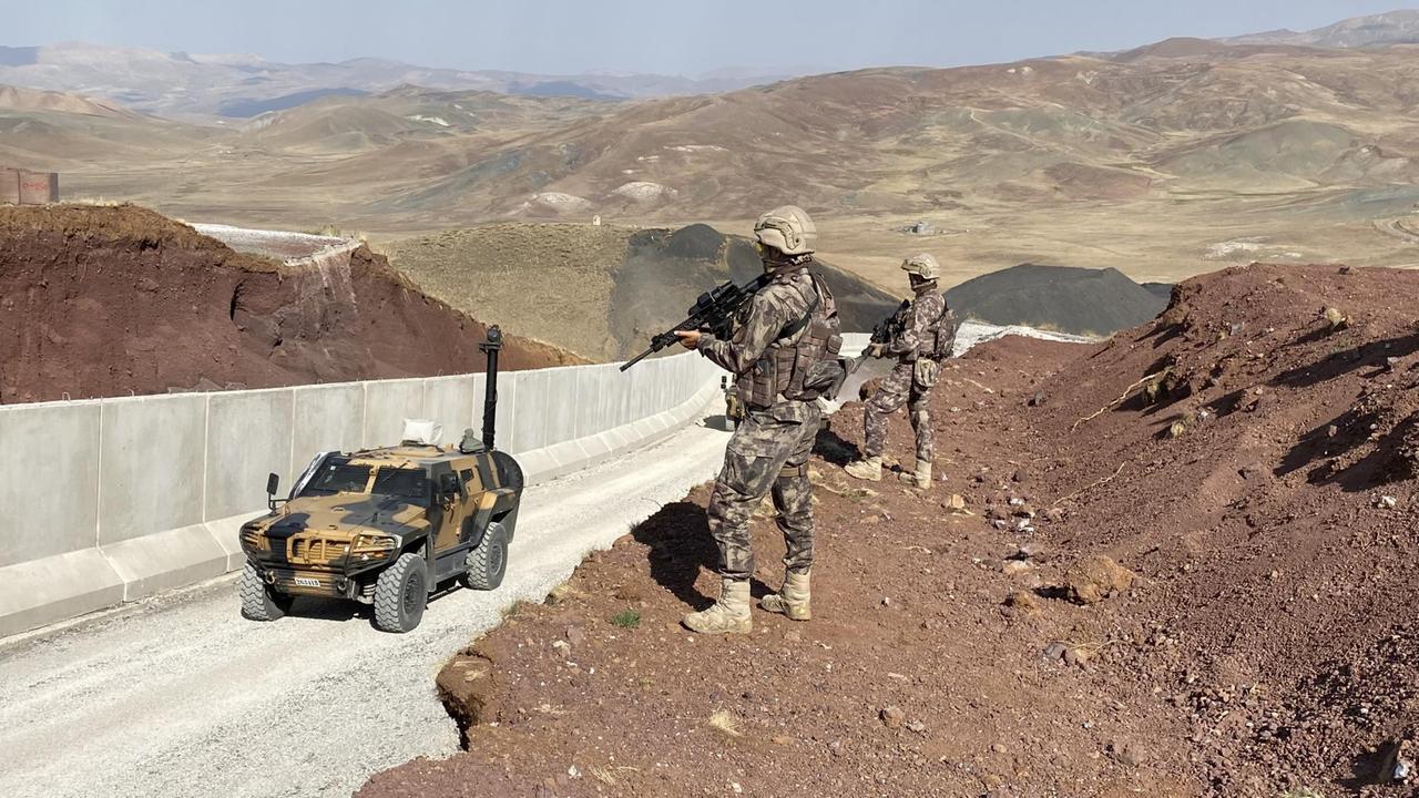 Ein Militärfahrzeug fährt eine Mauer entlang, von oben von bewaffneten Männern beobachtet.