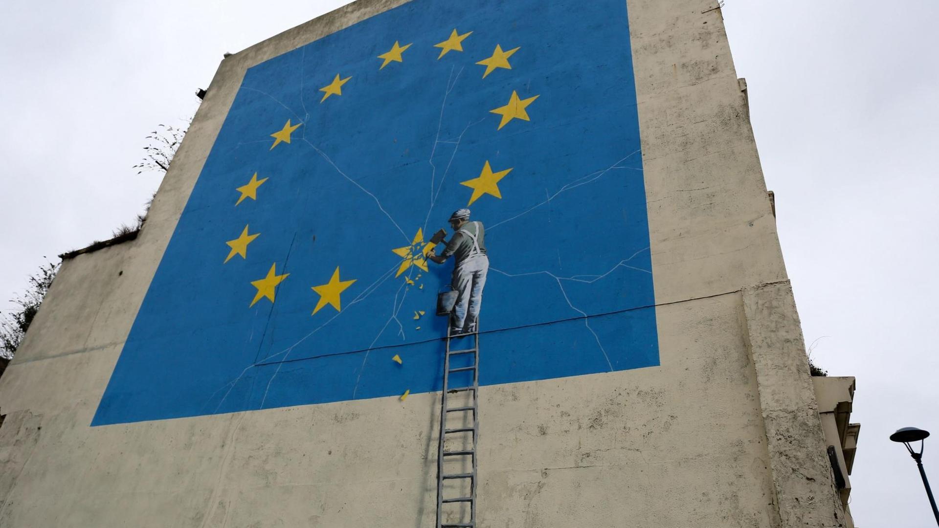 Das Foto zeigt das Wand-Bild von Banksy in der Stadt Dover. Ein Mann macht mit einem Hammer einen Stern in der Fahne von der EU kaputt.
