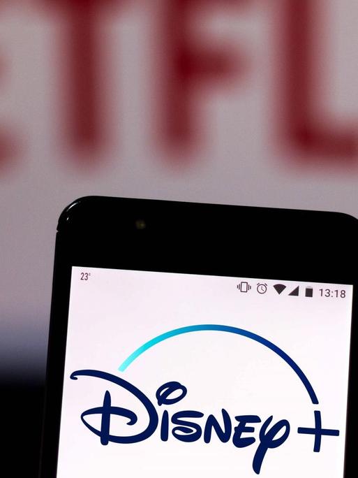 Auf einem Handy leuchtet das Logo von Disney+, im Hintergrund verschwommen das Netflix-Logo.