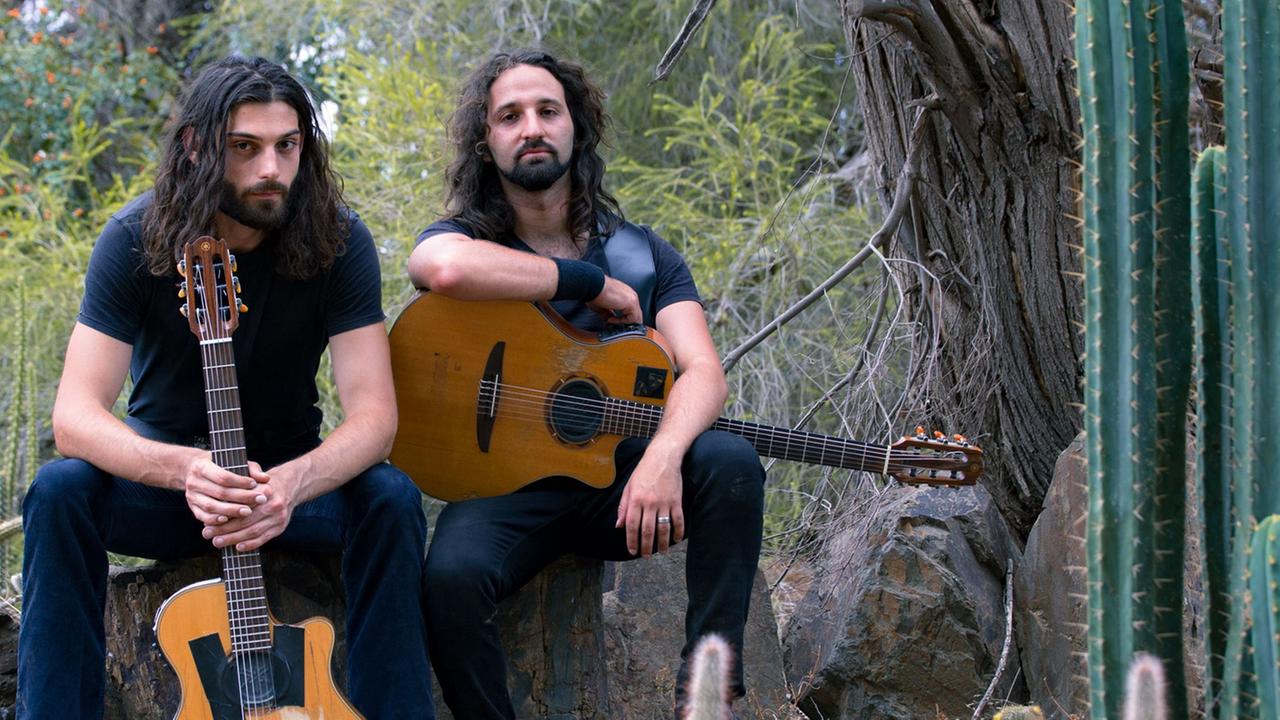 Zwei Männer mit langen, schwarzen Haaren und akustischen Gitarren sitzen auf einem Stein nebeneinander. Zwei Männer mit langen, schwarzen Haaren und akustischen Gitarren sitzen auf einem Stein nebeneinander.