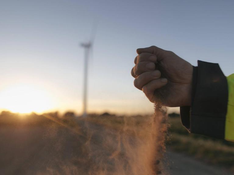 Ein Mann lässt Sand durch seine Hand rieseln, im Hintergrund ein Windrad auf einem Feld im Sonnenuntergang.