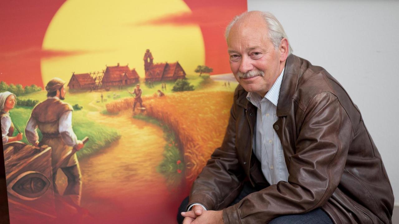 Klaus Teuber sitzt vor dem Bild seines Spiels "Die Siedler von Catan".