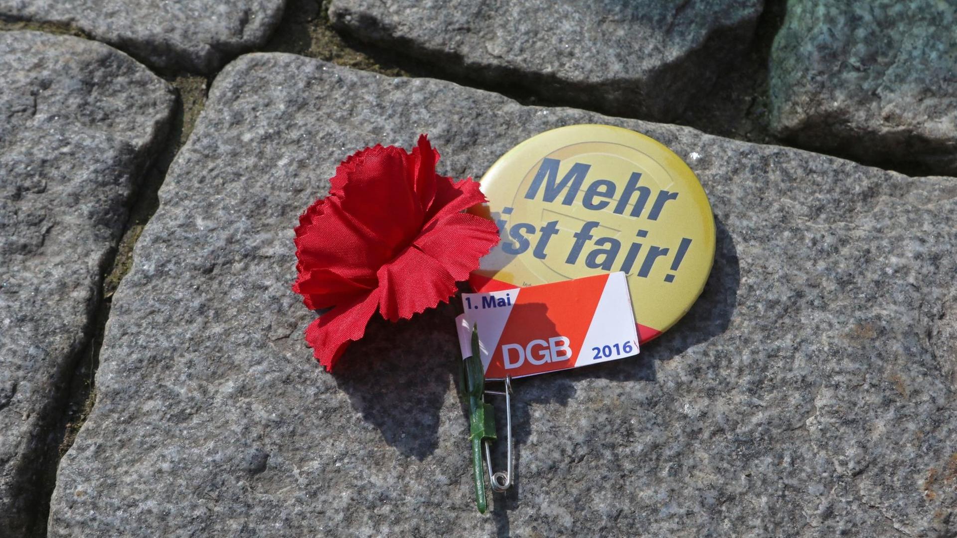 ILLUSTRATION - Ein Anstecker des Deutschen Gewerkschaftsbundes DGB mit einer Mai-Nelke und ein Sticker mit der Aufschrift "Mehr ist fair!" liegen am 01.05.2016 während einer DGB-Kundgebung auf dem Straßenpflaster des Marktplatzes in Wernigerode (Sachsen-Anhalt).