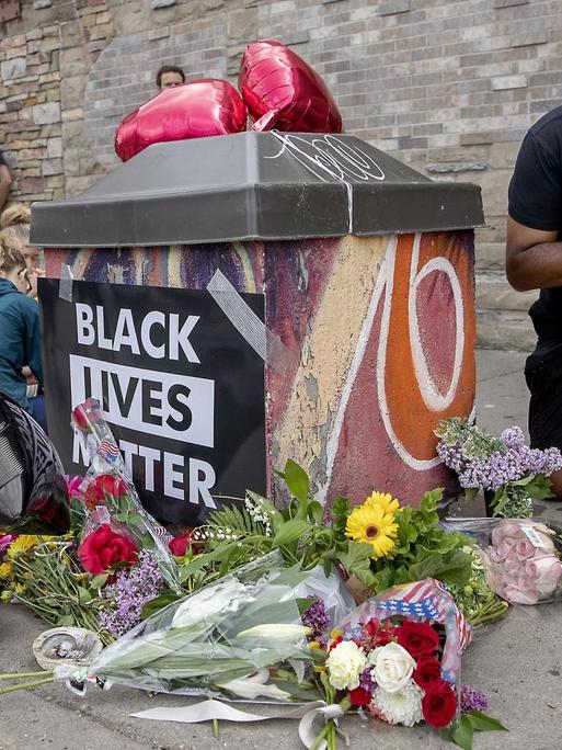 An einer Gedenkstelle mit Blumen und einem "Black Lives Matter"-Plakat trauern Menschen um George Floyd.