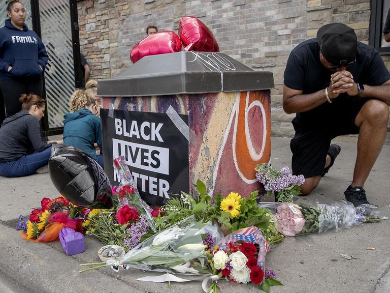 An einer Gedenkstelle mit Blumen und einem "Black Lives Matter"-Plakat trauern Menschen um George Floyd.