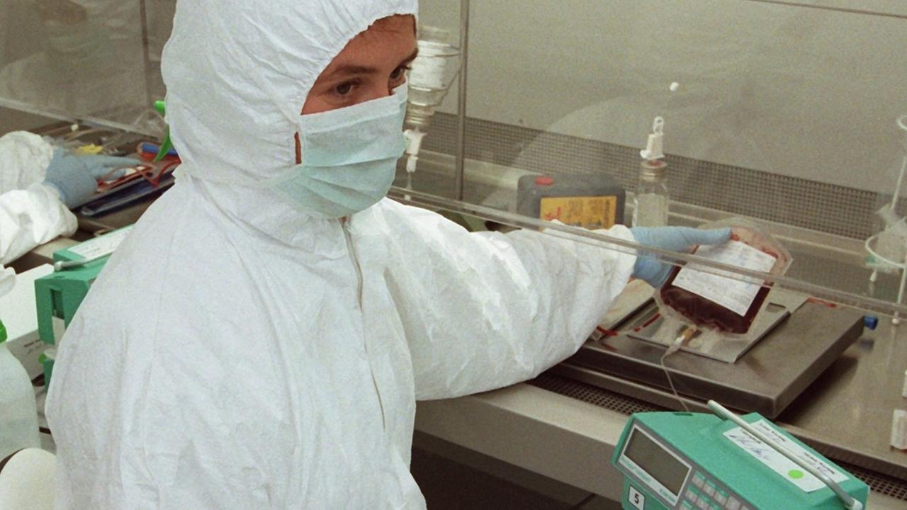 Eine Person in einem weißen Anzug sitzt mit Mundschutz in einem Labor und arbeitet mit einer Blutprobe