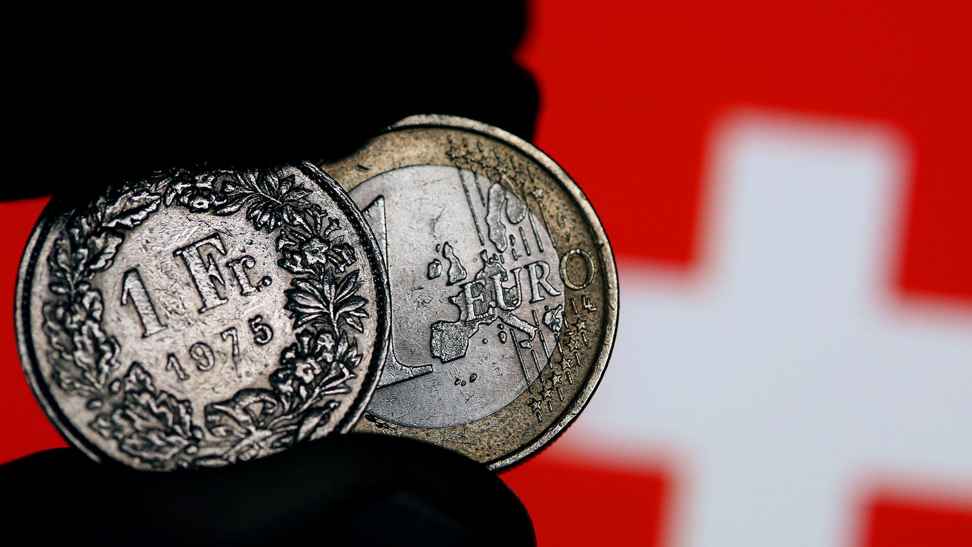 Eine Euro-Münze und ein Schweizer Franken werden am 15.01.2015 in Köln (Nordrhein-Westfalen) vor der Fahne der Eidgenossen zwischen den Fingern gehalten. Völlig überraschend hat die Schweizerische Nationalbank (SNB) am Donnerstag die Kopplung des Franken an den Euro aufgehoben und damit Turbulenzen an den Finanzmärkten ausgelöst.