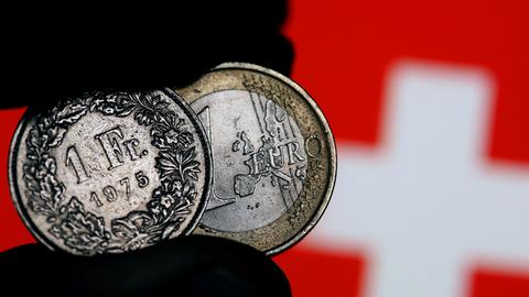 Eine Euro-Münze und ein Schweizer Franken werden am 15.01.2015 in Köln (Nordrhein-Westfalen) vor der Fahne der Eidgenossen zwischen den Fingern gehalten. Völlig überraschend hat die Schweizerische Nationalbank (SNB) am Donnerstag die Kopplung des Franken an den Euro aufgehoben und damit Turbulenzen an den Finanzmärkten ausgelöst.