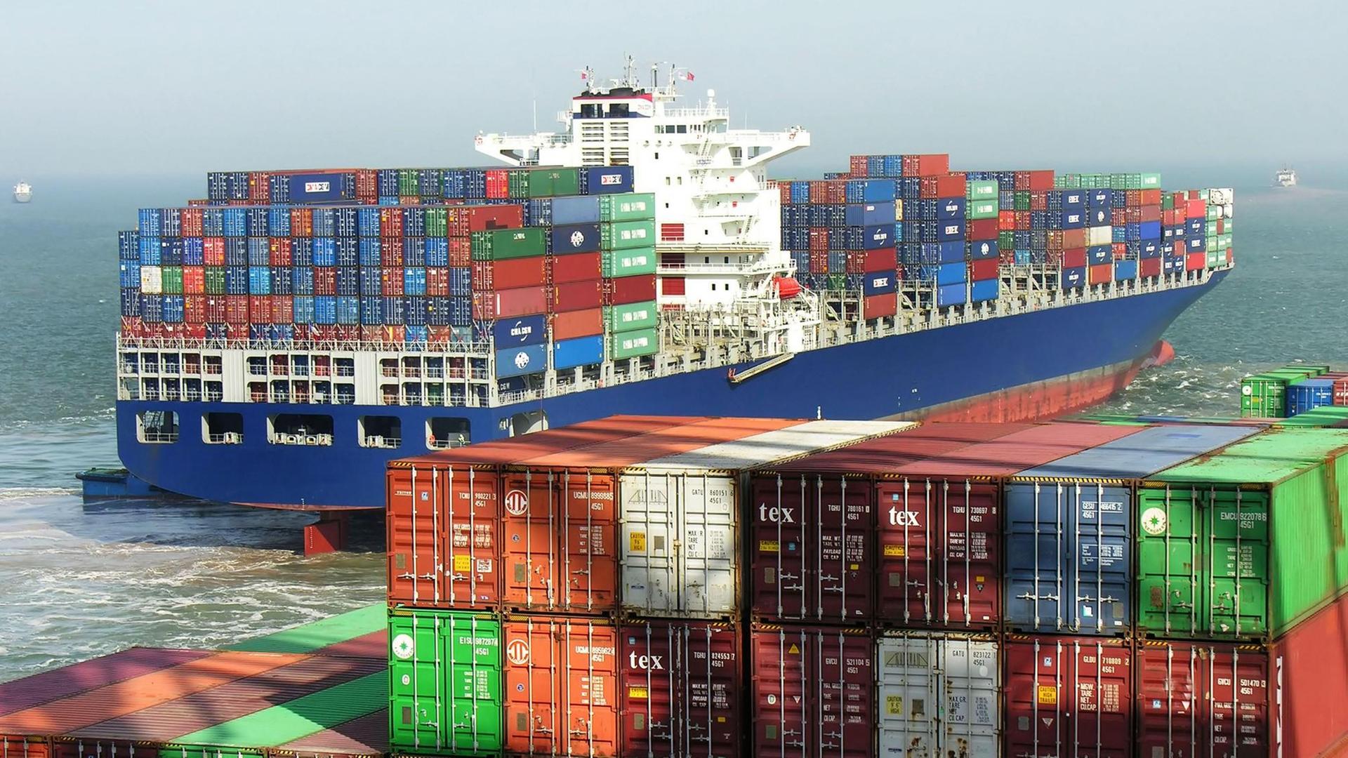 Ein mit vielen Container beladenes Schiff fährt auf dem Meer, im Vordergrund sind noch viele Container im Hafen zu sehen