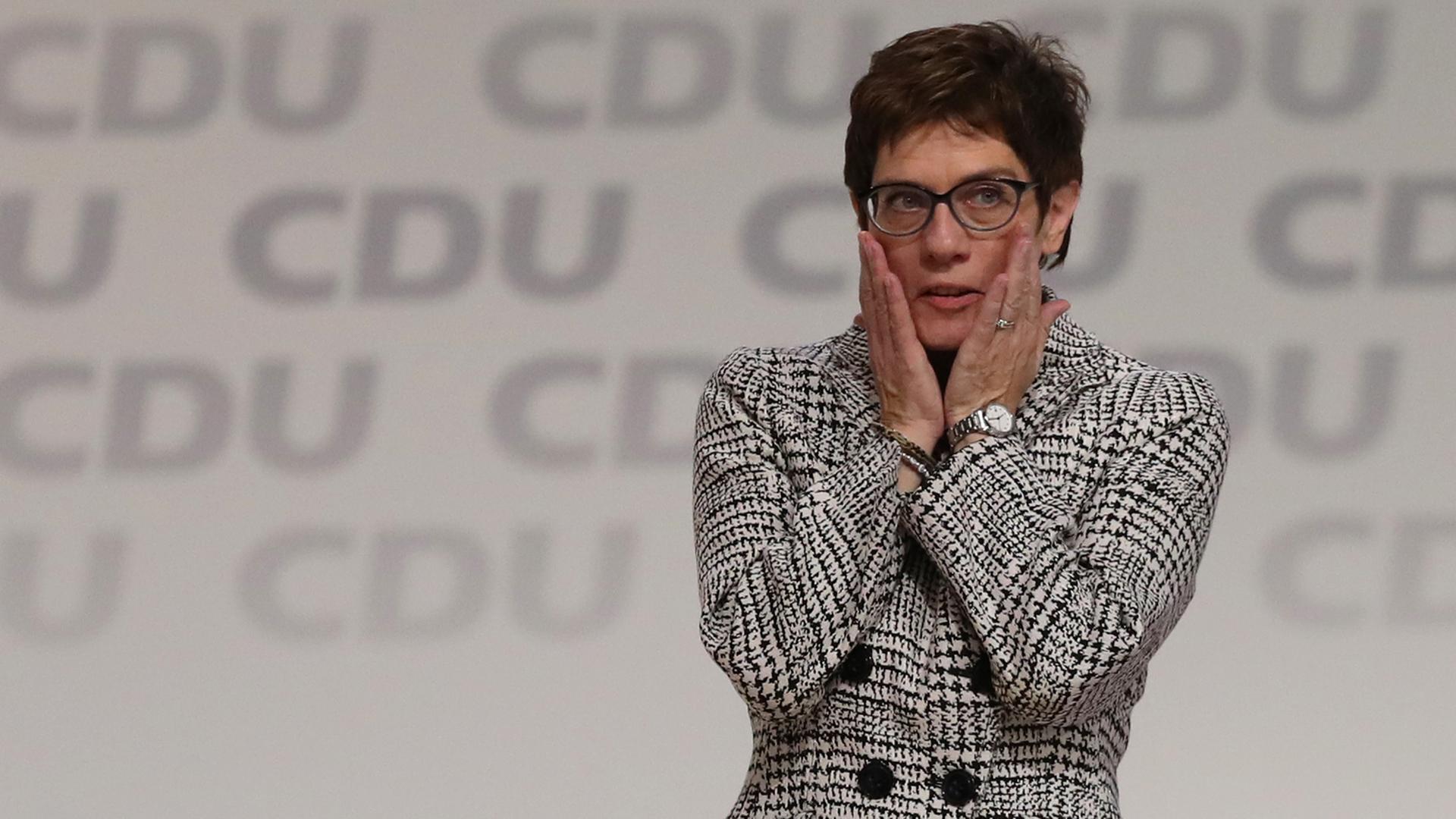 Annegret Kramp-Karrenbauer steht in einem schwarz-weiß-karrierten Sakko auf der Bühne, die Aufschrift "CDU" ist mehrfach hinter ihr zu sehen. Sie hält ihre Hände an ihre Wangen.